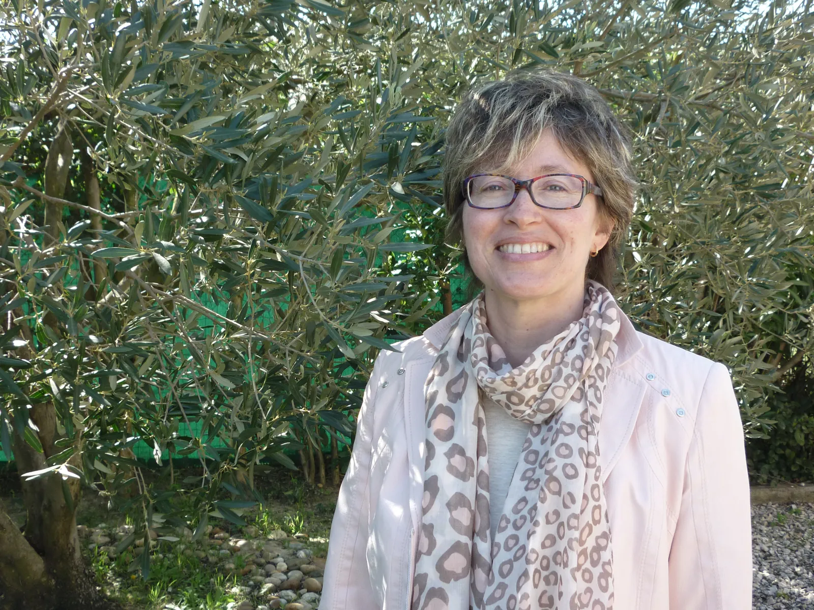 Véronique Férant souriante devant un olivier, en veste rose pâle et écharpe.